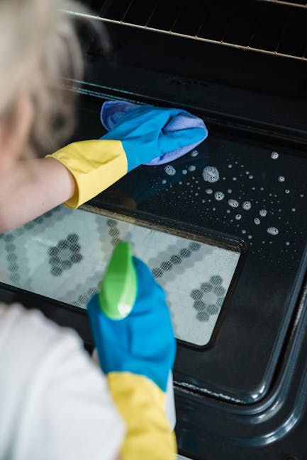 Close-up of a person wearing blue and yellow rubber gloves, cleaning the black glass door of a modern oven with a blue cloth, focusing on the surface which appears streak-free and slightly wet. Visibl