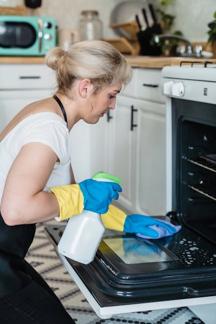 Close-up of a person wearing blue and yellow rubber gloves, cleaning the black glass door of a modern oven with a blue cloth, focusing on the surface which appears streak-free and slightly wet. Visibl