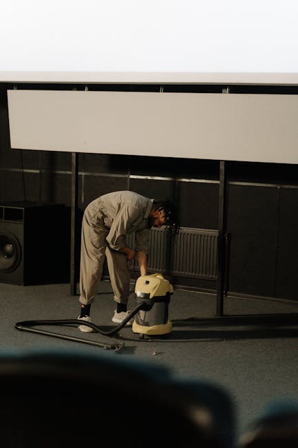 A professional cleaner dressed in a white protective suit, gloves, and shoe covers is using a wet vacuum or carpet extractor to perform deep cleaning and sanitisation of a beige carpet in a bright, mo